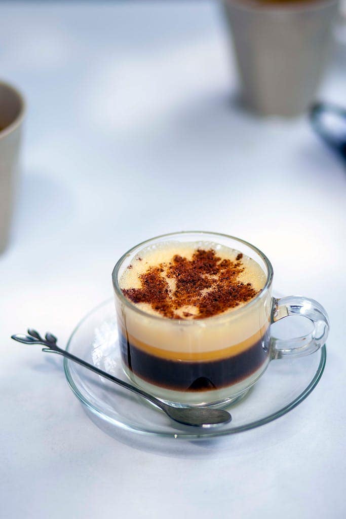 A close-up of a layered coffee in a glass cup with spoon on saucer, perfect for dessert lovers.